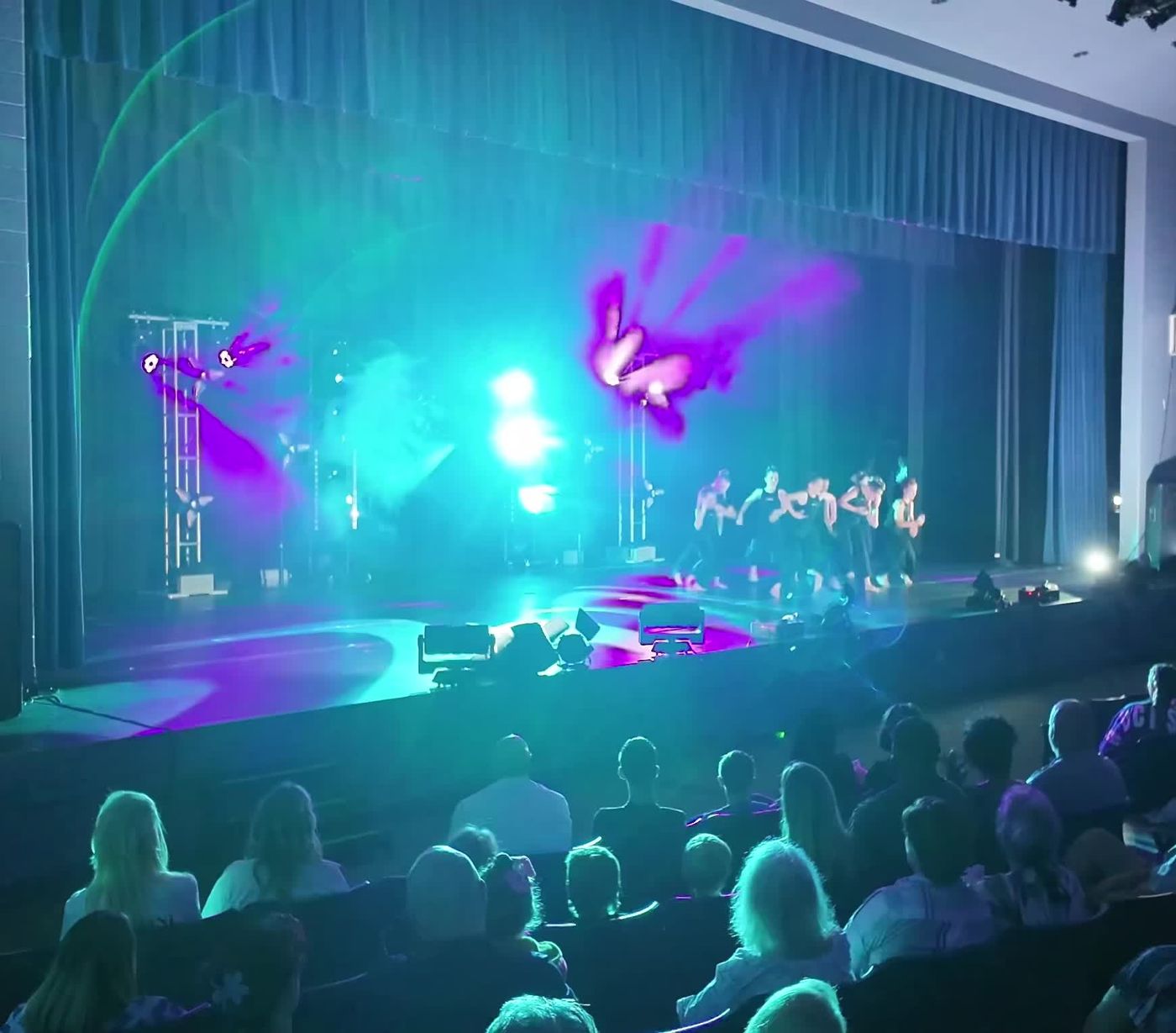 Dancers performing on stage under truss towers and magenta beams in front of a full audience
