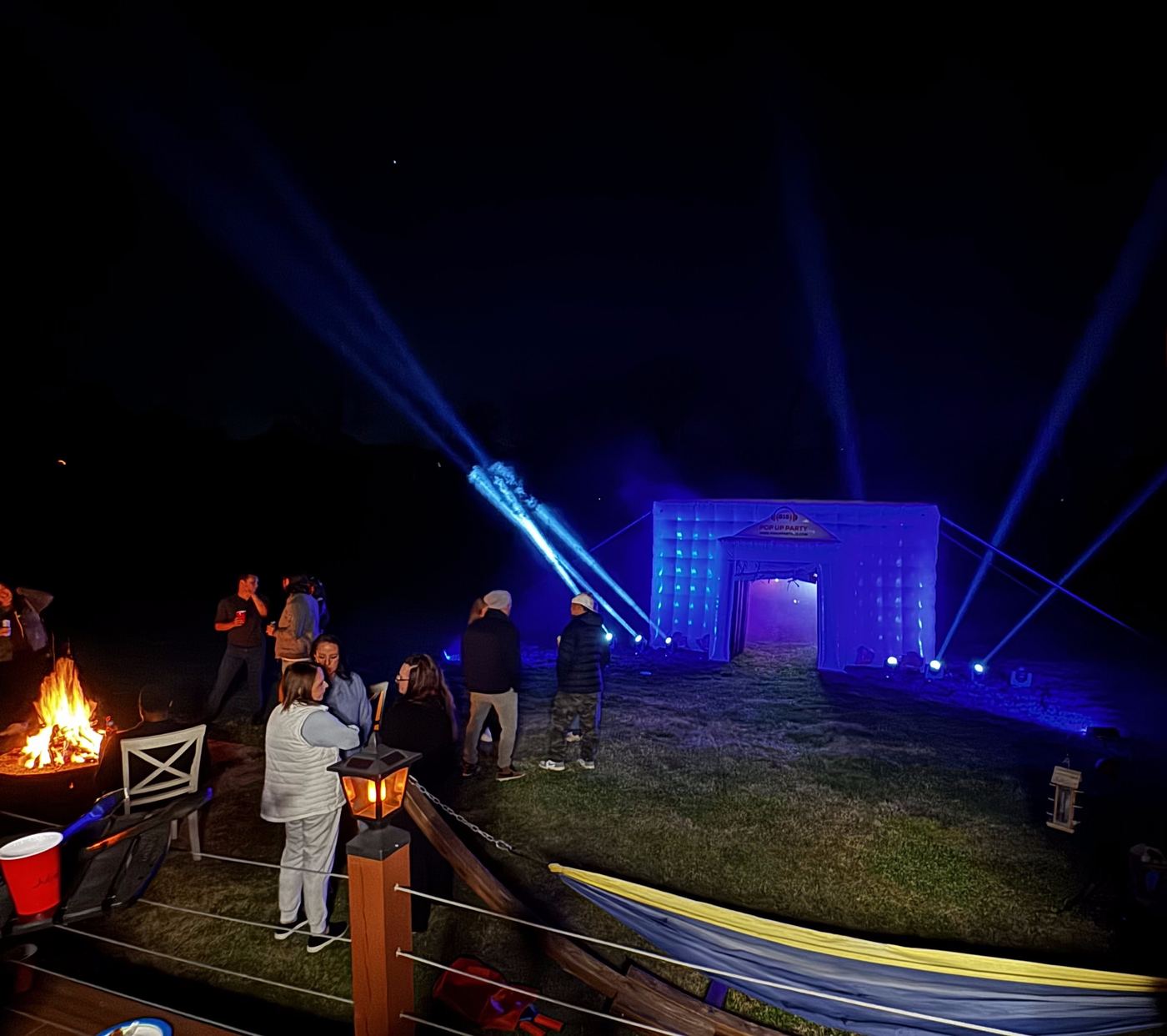 The pop-up party tent glowing blue at night, beams overhead, guests around a fire pit